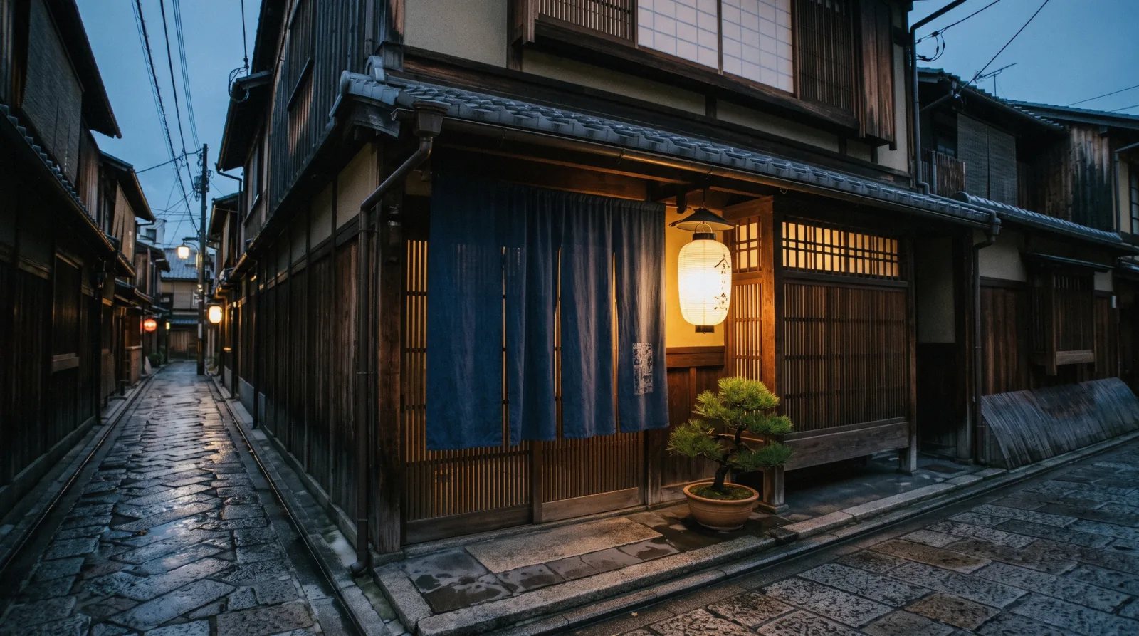 A restored machiya inn frontage with a noren curtain and lantern at dusk, where to stay in Kyoto Japan
